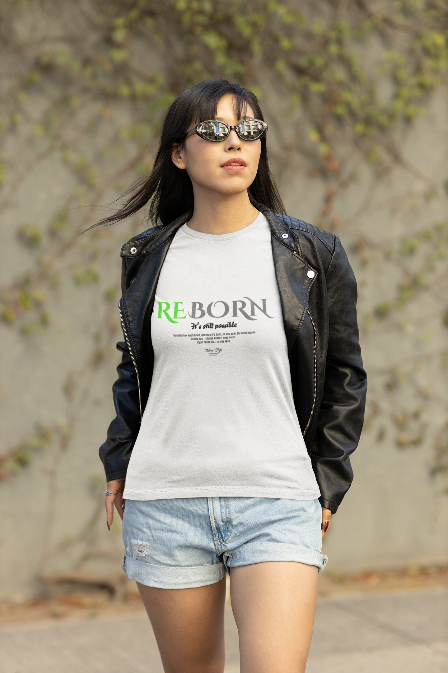 Female model in black leather jacket wearing the REBORN t-shirt in white. Walking with confidence, wind in her hair, sunglasses on. The green-accented “It’s still possible” message stands out boldly against the clean white fabric.