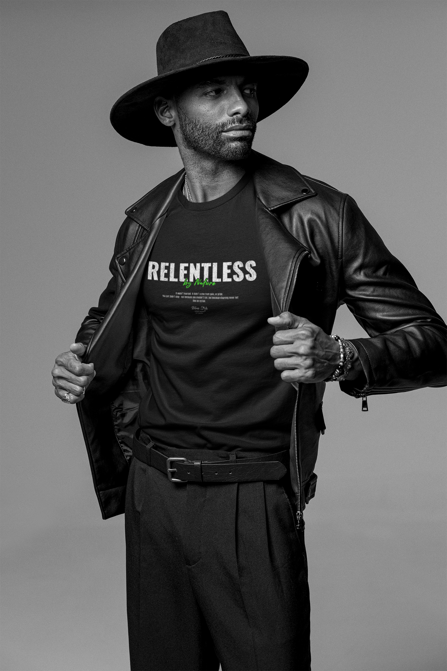 A confident man in a wide-brimmed hat and leather jacket reveals the RELENTLESS t-shirt underneath. The shirt features bold, textured typography with the phrase “By Nature” in green, embodying the quiet power and instinct-driven force of the design.