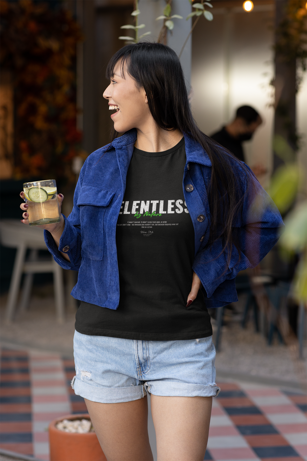A cheerful woman in a blue corduroy jacket and denim shorts enjoys a drink outdoors while wearing the RELENTLESS t-shirt. The bold text 'By Nature' pops in green beneath the main word, reinforcing the shirt’s raw, unstoppable energy.