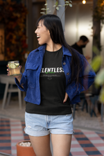 A cheerful woman in a blue corduroy jacket and denim shorts enjoys a drink outdoors while wearing the RELENTLESS t-shirt. The bold text 'By Nature' pops in green beneath the main word, reinforcing the shirt’s raw, unstoppable energy.