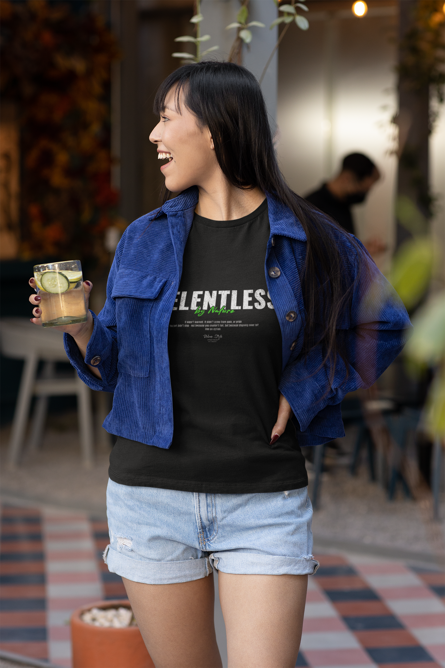 A cheerful woman in a blue corduroy jacket and denim shorts enjoys a drink outdoors while wearing the RELENTLESS t-shirt. The bold text 'By Nature' pops in green beneath the main word, reinforcing the shirt’s raw, unstoppable energy.