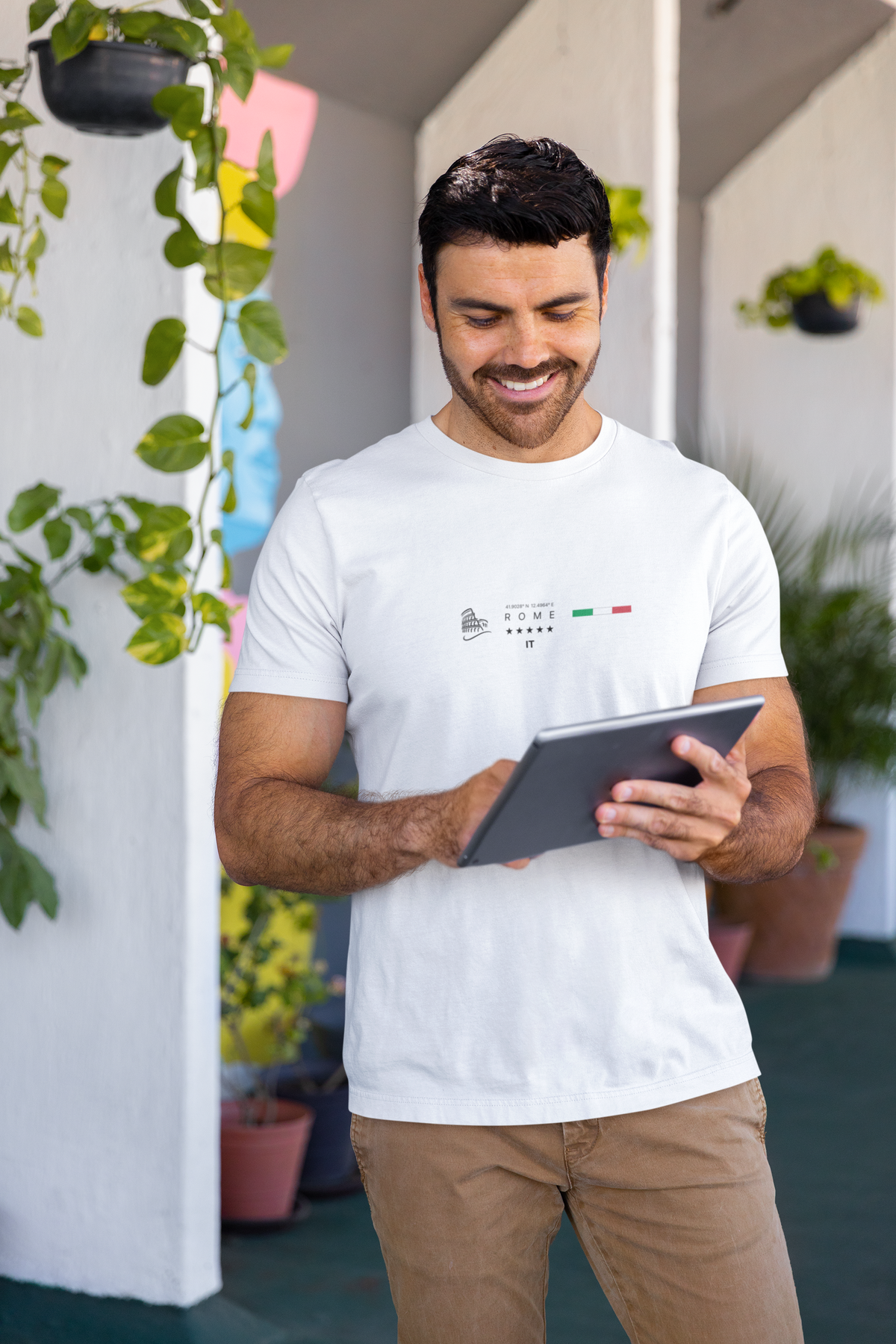 A smiling man standing on a bright balcony, wearing a white "ROME" t-shirt featuring a minimal Colosseum icon, 5 stars, and the Italian flag. He holds a tablet while surrounded by potted plants and sunlight, creating a relaxed and modern lifestyle vibe.