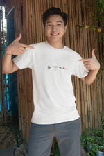 A cheerful man pointing at the "ROME" t-shirt he’s wearing, emphasizing the design with both hands. The shirt shows the Colosseum, 5 stars, and the Italian flag. He stands against a bamboo wall with plants, in a casual outdoor setting filled with natural light and a playful tone.