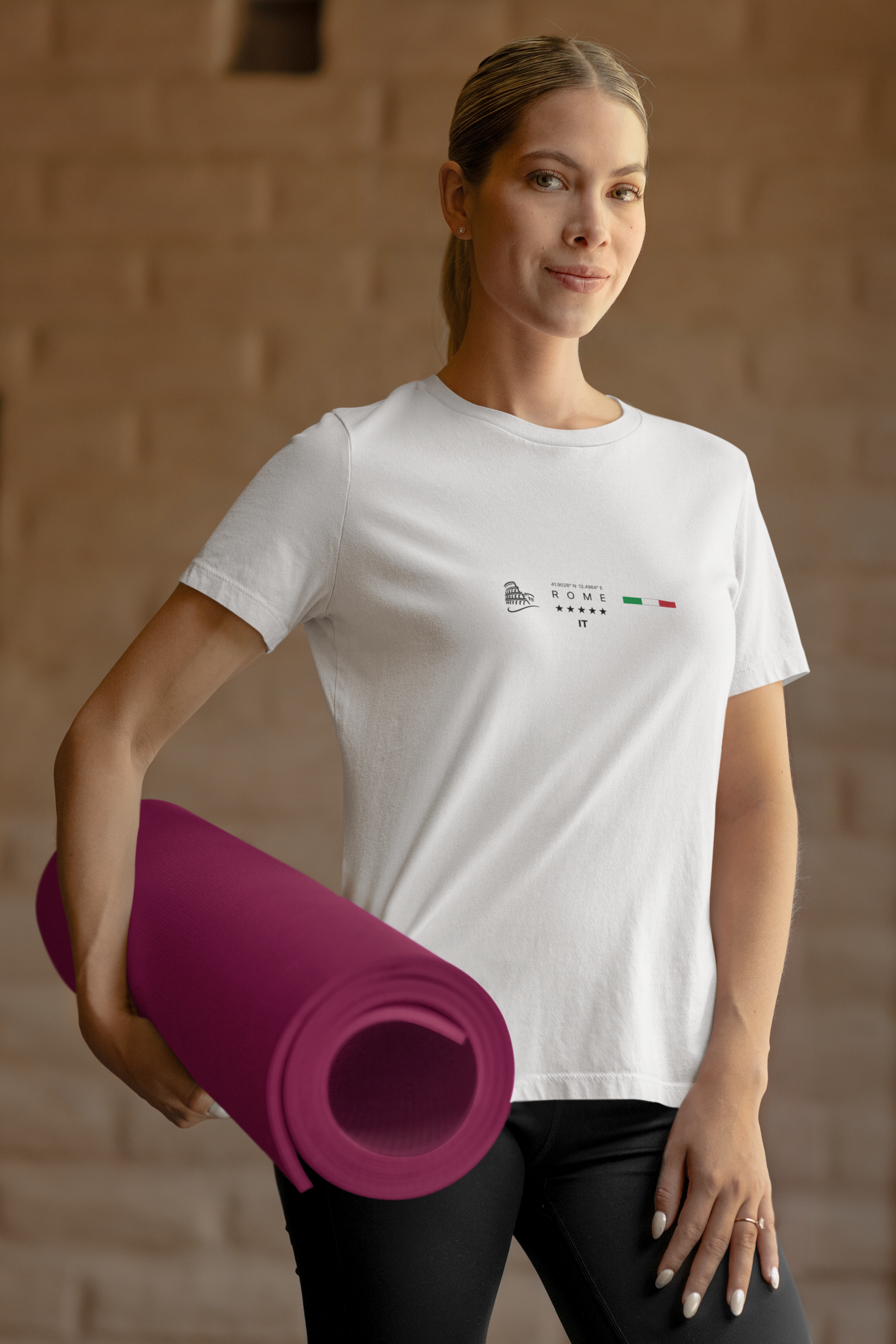 A confident woman holding a rolled yoga mat in one hand, wearing a white "ROME" t-shirt with the Colosseum illustration and Italian flag. Set indoors with warm lighting and a soft brick wall background, her pose and athletic style highlight calm strength and daily routine.