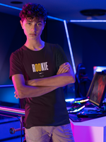 Young male model standing confidently in a neon-lit gaming room, wearing the “ROOKIE” t-shirt in black. The design features bold white text with the letters “OO” in yellow. His arms are crossed, and the background includes gaming monitors and blue-violet LED accents.
