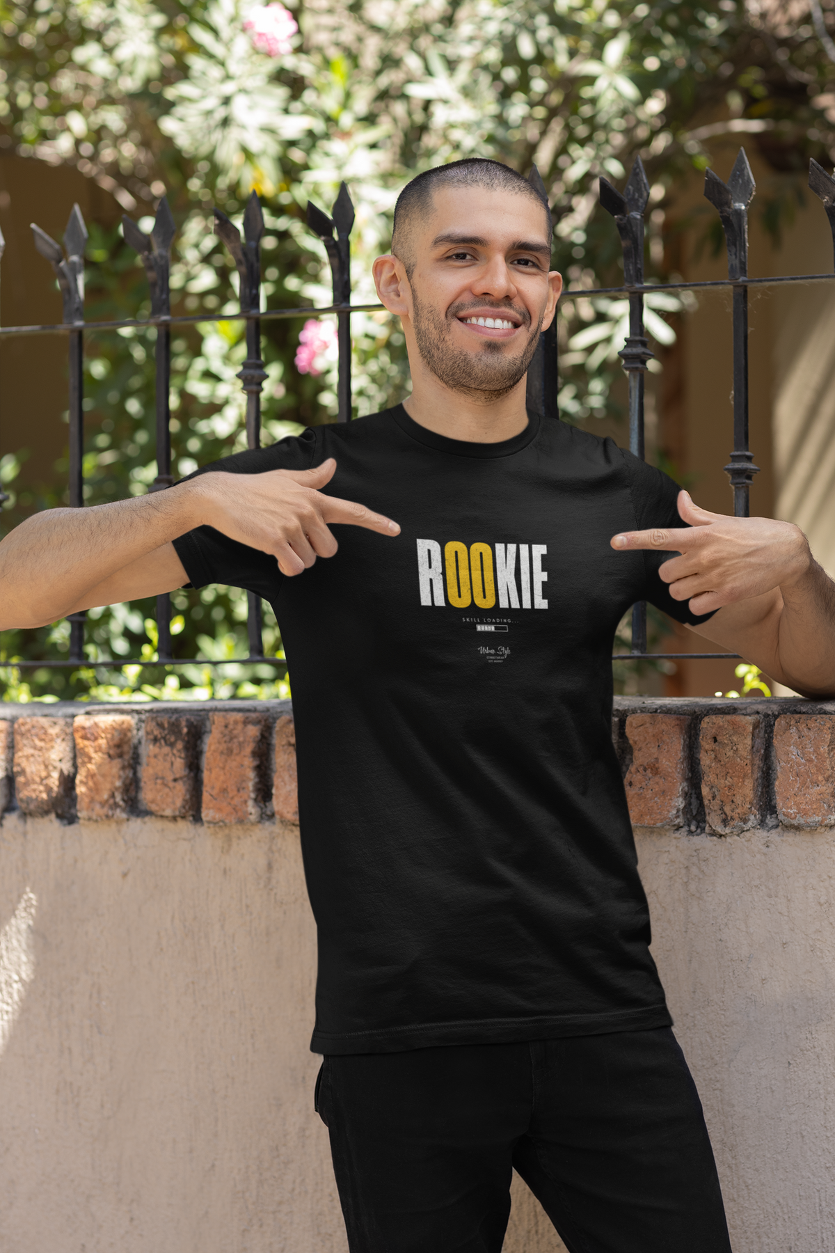 Male model smiling confidently outdoors in front of a black iron fence, wearing the “ROOKIE” t-shirt in black. He’s pointing at the text design on the shirt, which features bold white letters with the “OO” in yellow. Sunlight and soft greenery complete the background, adding a casual and fresh vibe.