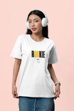 Female model standing against a soft pink background, wearing headphones and the “ROOKIE” t-shirt in white. The shirt features bold black text with the “OO” in yellow. She holds the hem of her shirt casually while giving a confident, stylish look. Denim skirt and smart accessories complete the modern, youthful vibe.