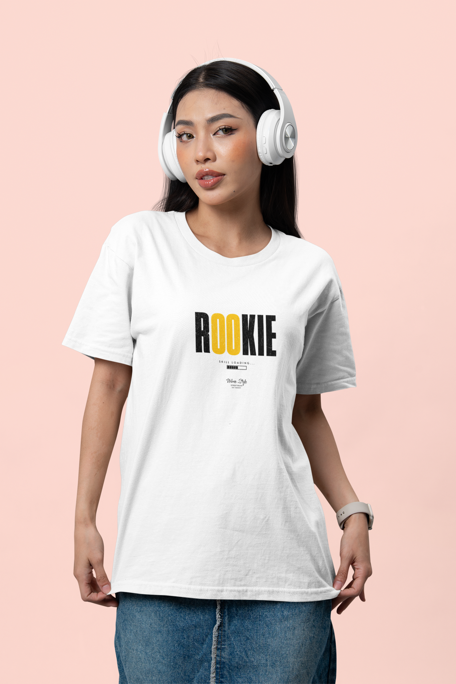 Female model standing against a soft pink background, wearing headphones and the “ROOKIE” t-shirt in white. The shirt features bold black text with the “OO” in yellow. She holds the hem of her shirt casually while giving a confident, stylish look. Denim skirt and smart accessories complete the modern, youthful vibe.
