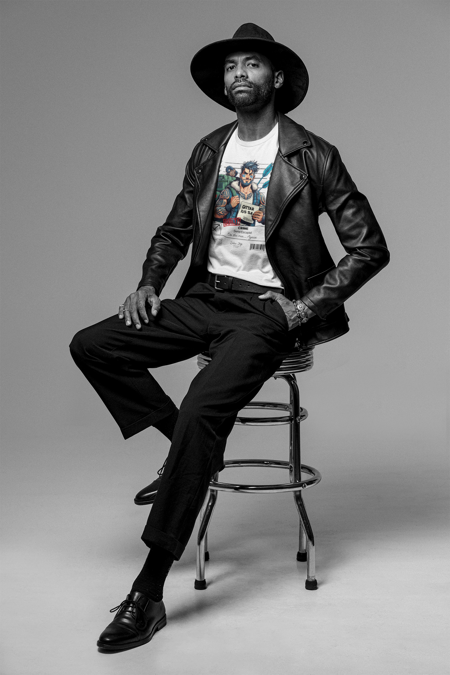 White t-shirt worn by a stylish man seated on a chrome stool in a studio. He pairs the Sagittarius “Serial Escapist” graphic with a wide-brim hat, leather jacket, and formal pants. Monochrome setting contrasts with the vibrant full-color shirt design.