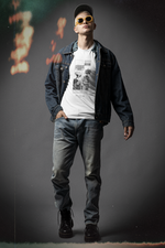 A man poses confidently against a moody gray backdrop, wearing a white graphic tee layered under a denim jacket and paired with light-washed jeans. The shirt features a two-panel comic where an alien asks, “Do you feel any guilt after scratching the furniture?” and a smug cat replies, “I feel nothing. Except satisfiction.” His yellow sunglasses and rugged boots add a bold, urban edge to the overall look. Faint red and orange light leaks on the image add a vintage, analog vibe.