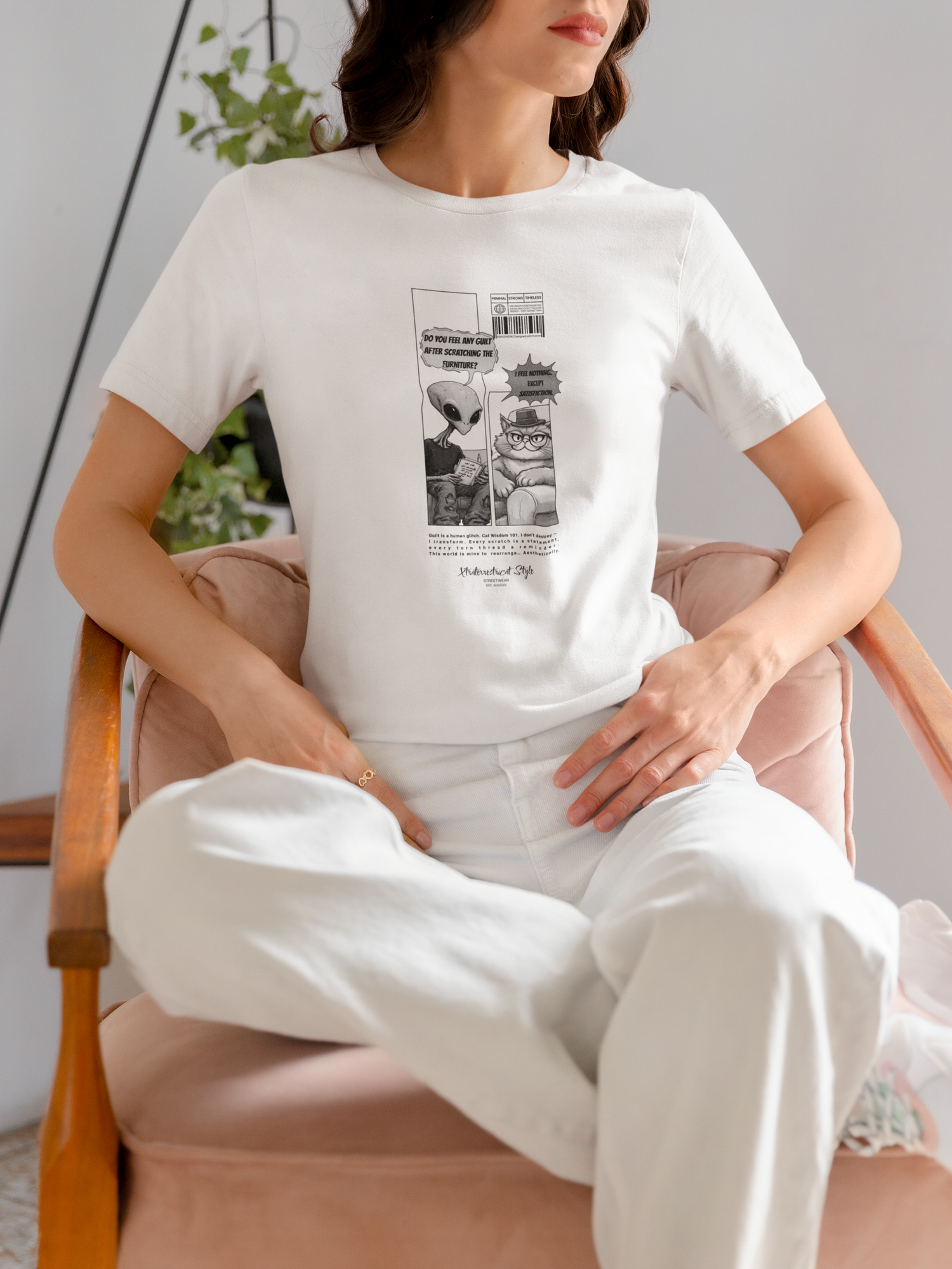 A woman sits relaxed on a pale pink armchair, wearing a white graphic t-shirt with a two-panel comic design. The left panel shows an alien asking, “Do you feel any guilt after scratching the furniture?” and the right panel features a smug cat in a fedora responding, “I feel nothing. Except satisfiction.” The woman pairs the shirt with white jeans, blending softly into the bright, minimalist setting. A leafy green plant peeks from the background, adding a touch of nature to the calm, stylish scene.