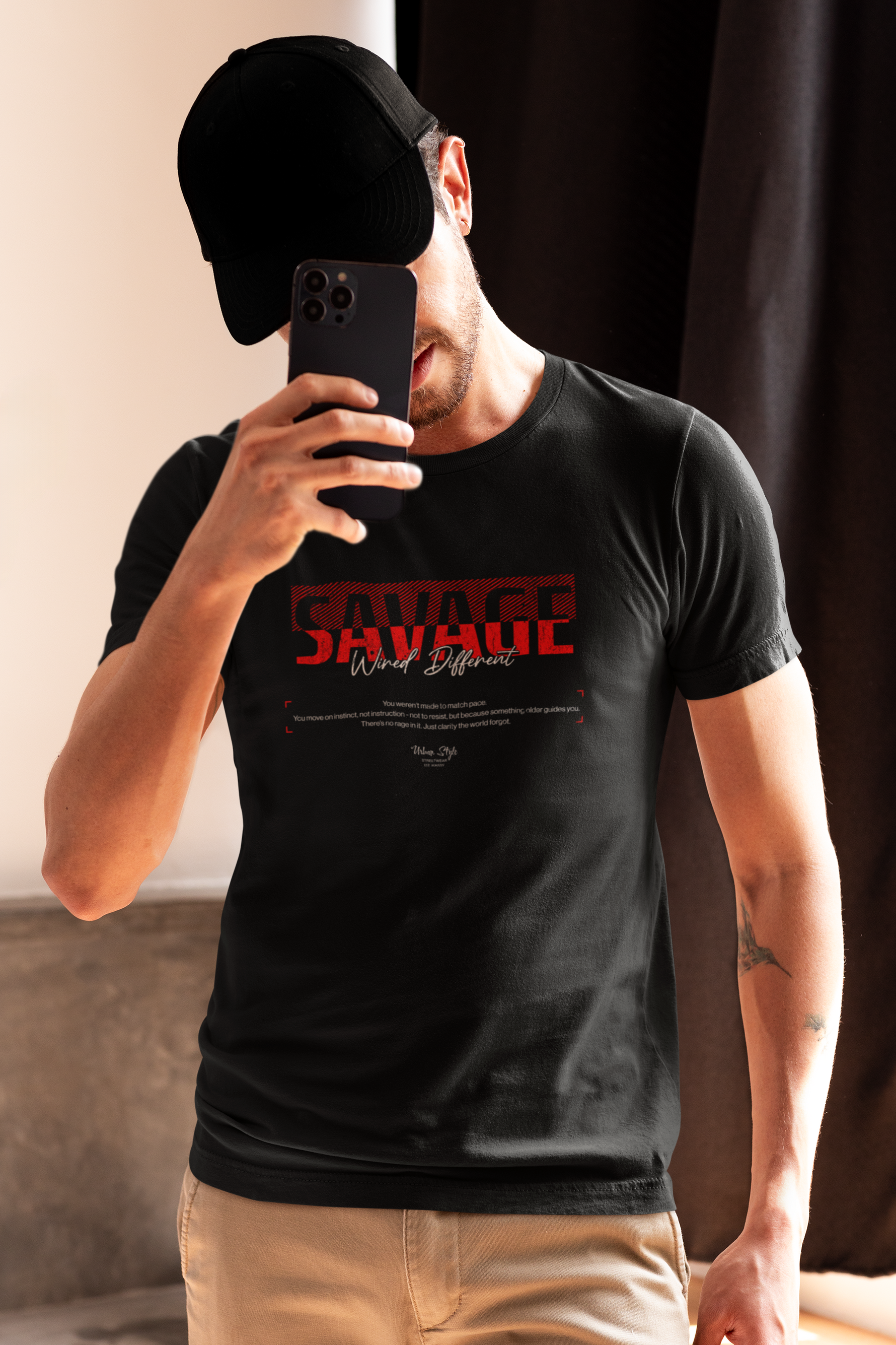 Man in beige pants and black cap taking a mirror selfie, wearing the black SAVAGE t-shirt with red “Wired Different” print — casual, confident, and unfiltered street energy.