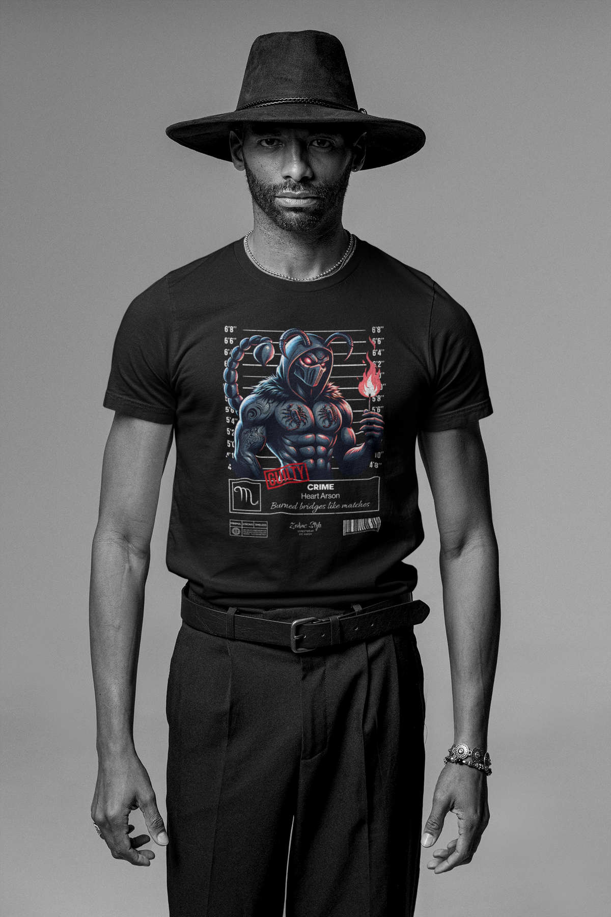 Black t-shirt worn by a man with a wide-brimmed hat, standing confidently in a studio setting. The design features a masked scorpion warrior holding a flame with a mugshot backdrop.