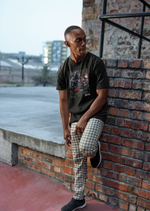 Black t-shirt worn by a man leaning against a brick wall outdoors. The mugshot-style design features a stylized scorpion warrior with a flame in hand, appearing fierce and bold.