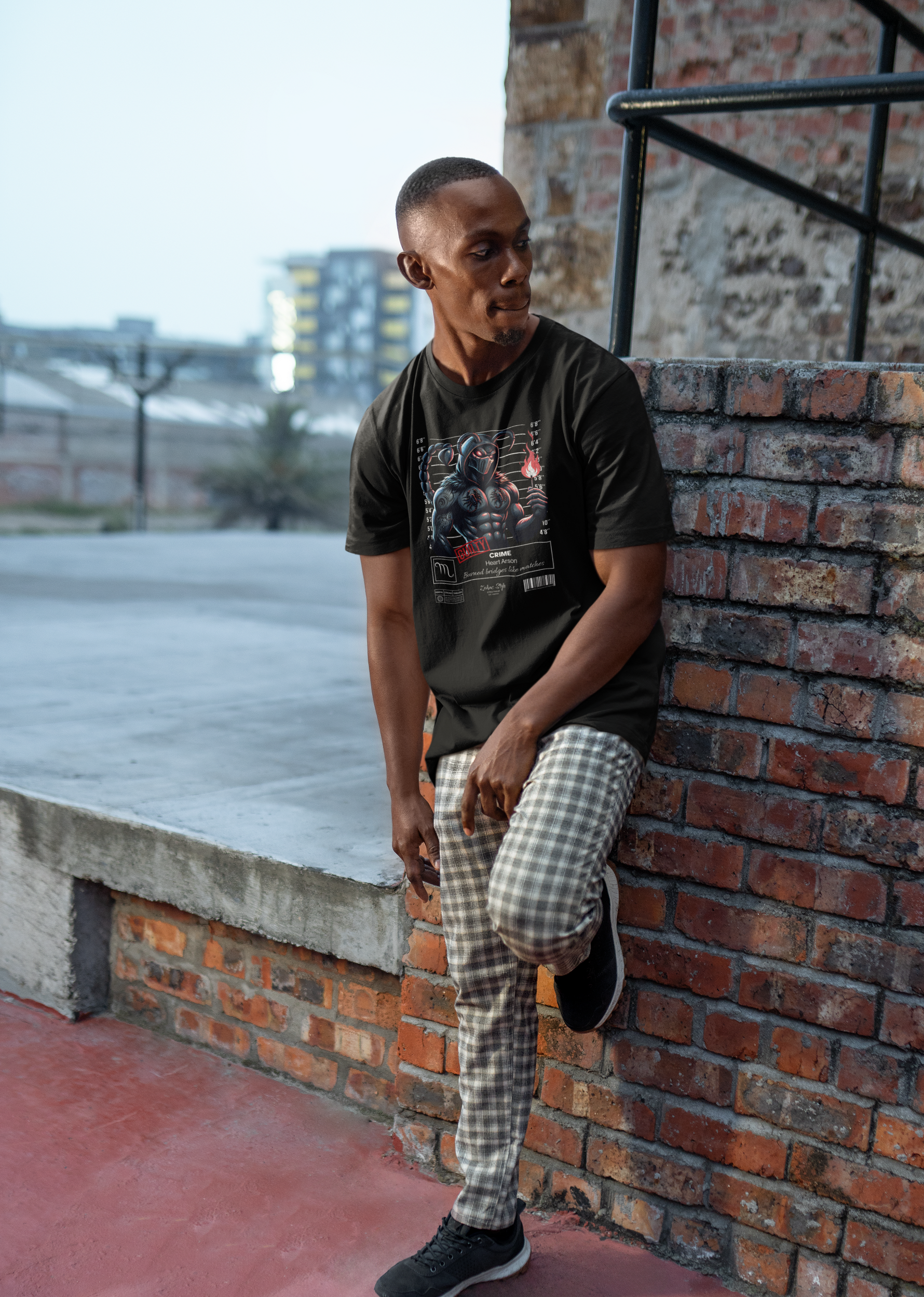 Black t-shirt worn by a man leaning against a brick wall outdoors. The mugshot-style design features a stylized scorpion warrior with a flame in hand, appearing fierce and bold.