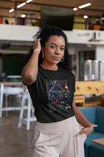 Black t-shirt worn by a woman indoors, gently holding her hair. She's standing in a cozy café. The design shows a muscular scorpion figure with tribal tattoos and a fiery torch.