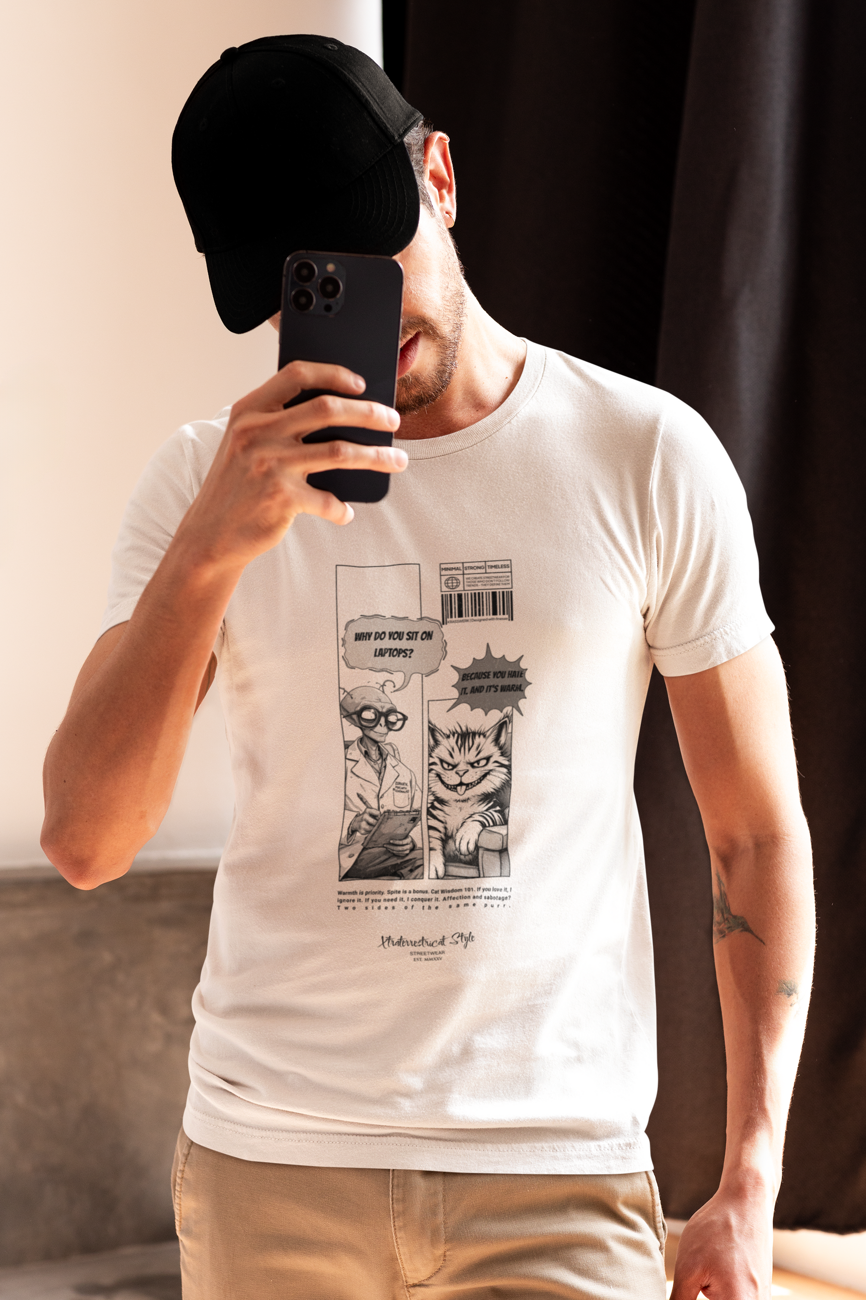 A man takes a mirror selfie in natural light, wearing a white t-shirt with a quirky alien-cat comic. The alien asks, “Why do you sit on laptops?” and the cat replies smugly, “Because you hate it. And it’s warm.” His face is partially hidden behind a phone, adding a casual, spontaneous vibe to the image.