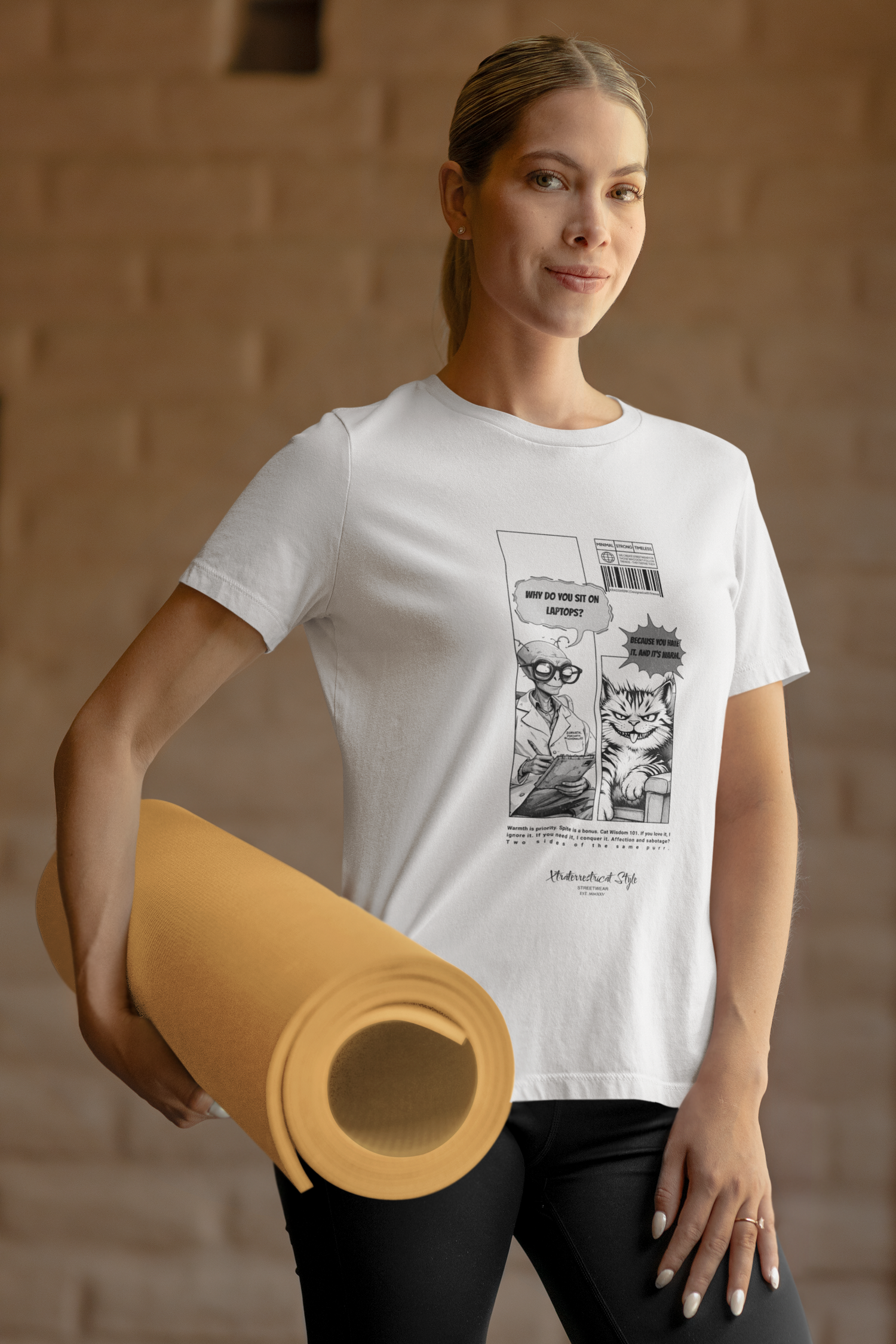 A woman stands confidently in a warm-toned studio, holding a rolled yoga mat in one hand. She wears a white t-shirt printed with a black-and-white comic design. The comic shows an alien asking, “Why do you sit on laptops?” and a smug-looking cat replying, “Because you hate it. And it's warm.” The overall vibe is modern and humorous, blending everyday fitness style with nerdy cat attitude.
