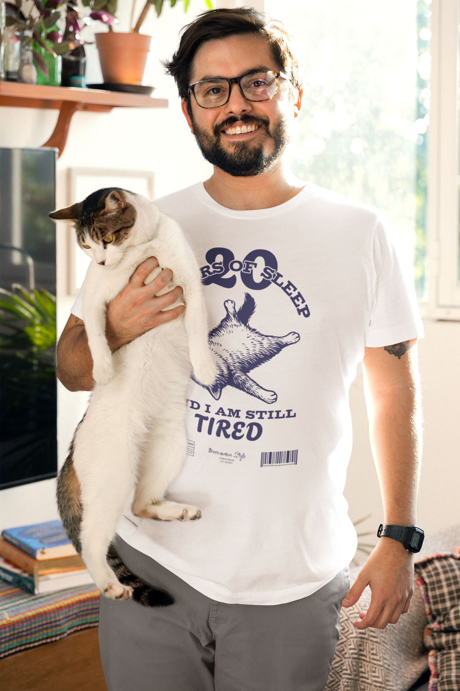 Smiling man holding a sleepy white and brown cat in one arm stands in a cozy, sunlit room. He wears a white t-shirt with a humorous design of a belly-up cat and the text: “20 HOURS OF SLEEP AND I AM STILL TIRED.”