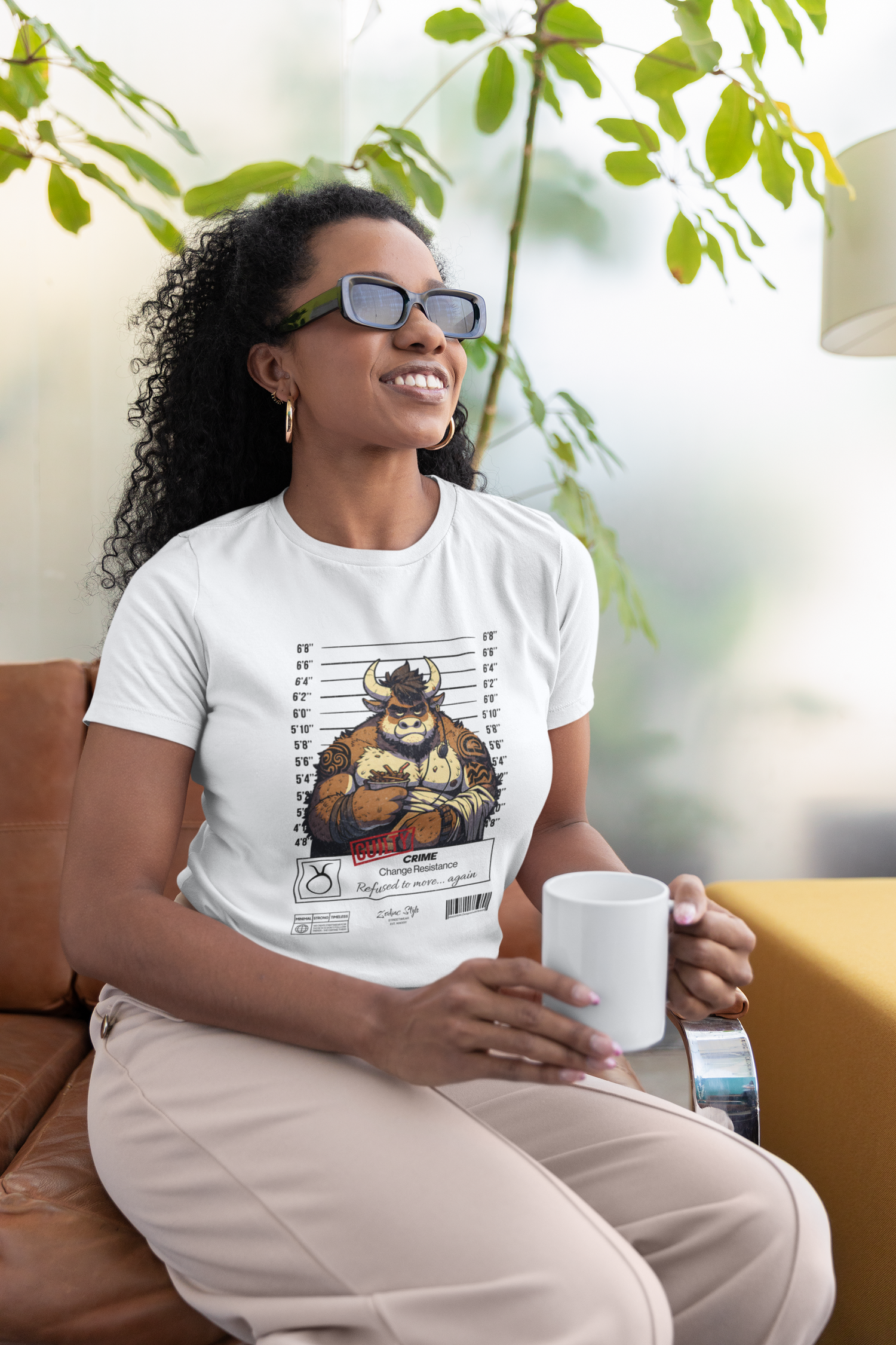 Indoor lifestyle setting — woman lounges with a coffee mug, wearing the TAURUS T-shirt in white. The design shows a tattooed bull in a mugshot with crossed arms. Below reads: “CRIME: Change Resistance. Refused to move… again.” Calm exterior, stubborn core — this one’s built to stay.