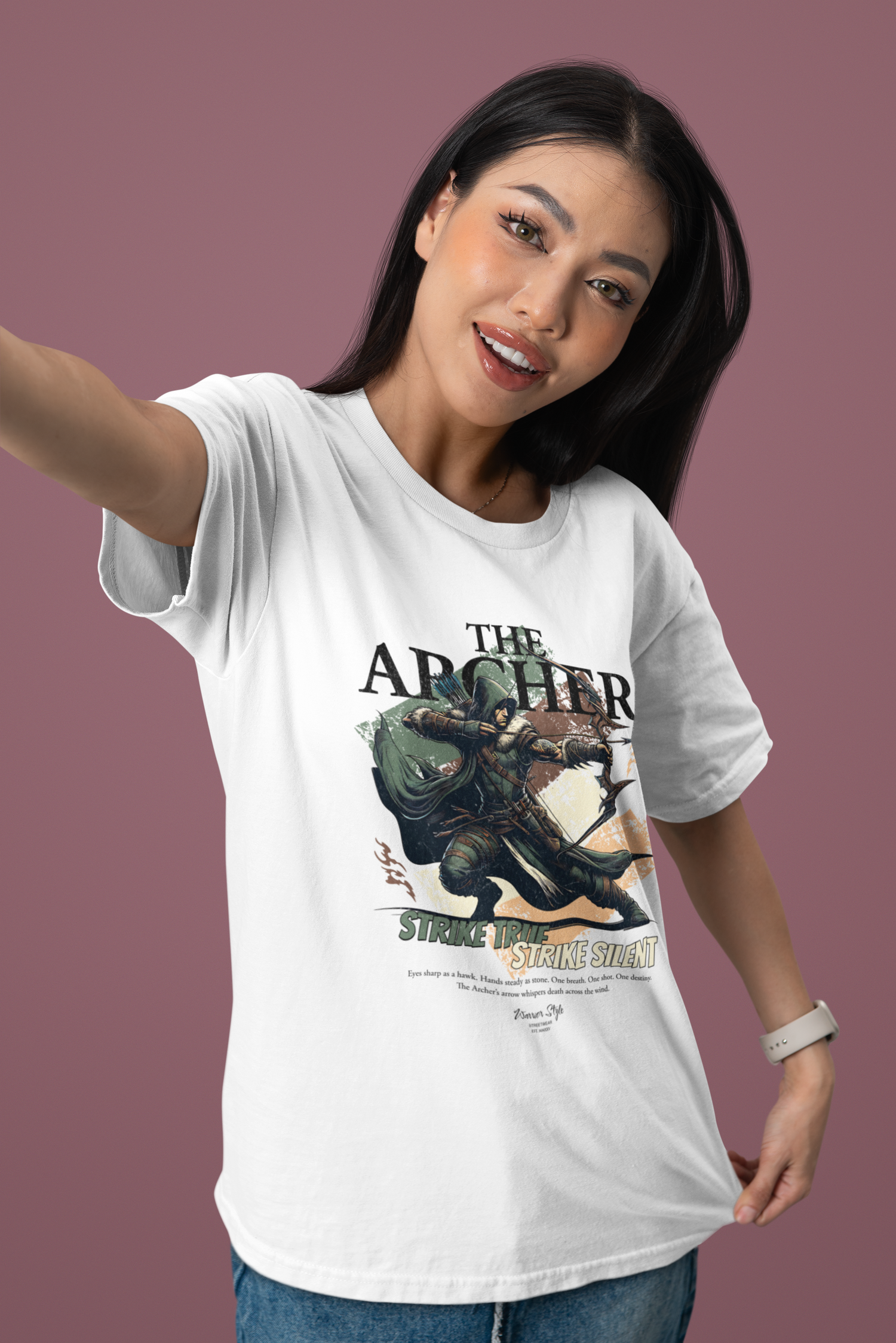 A confident woman smiling playfully while taking a selfie, wearing THE ARCHER t-shirt in white. The design features a hooded archer mid-draw with the slogan “Strike True. Strike Silent.” A soft rose background adds a fresh, modern touch that contrasts the bold artwork.