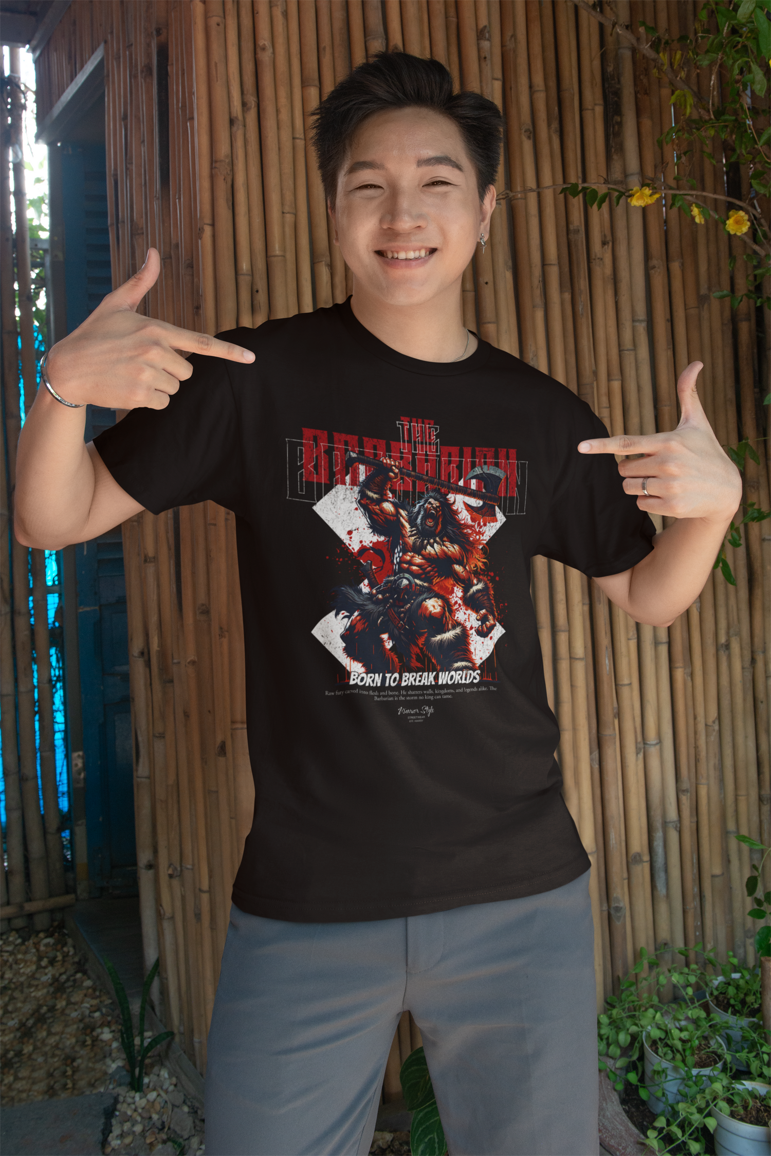 A man smiling and pointing at his chest while wearing the KRASSWERK 'THE BARBARIAN' graphic t-shirt in black. He stands in front of a rustic bamboo wall with warm daylight and plants around, radiating confidence and raw joy.