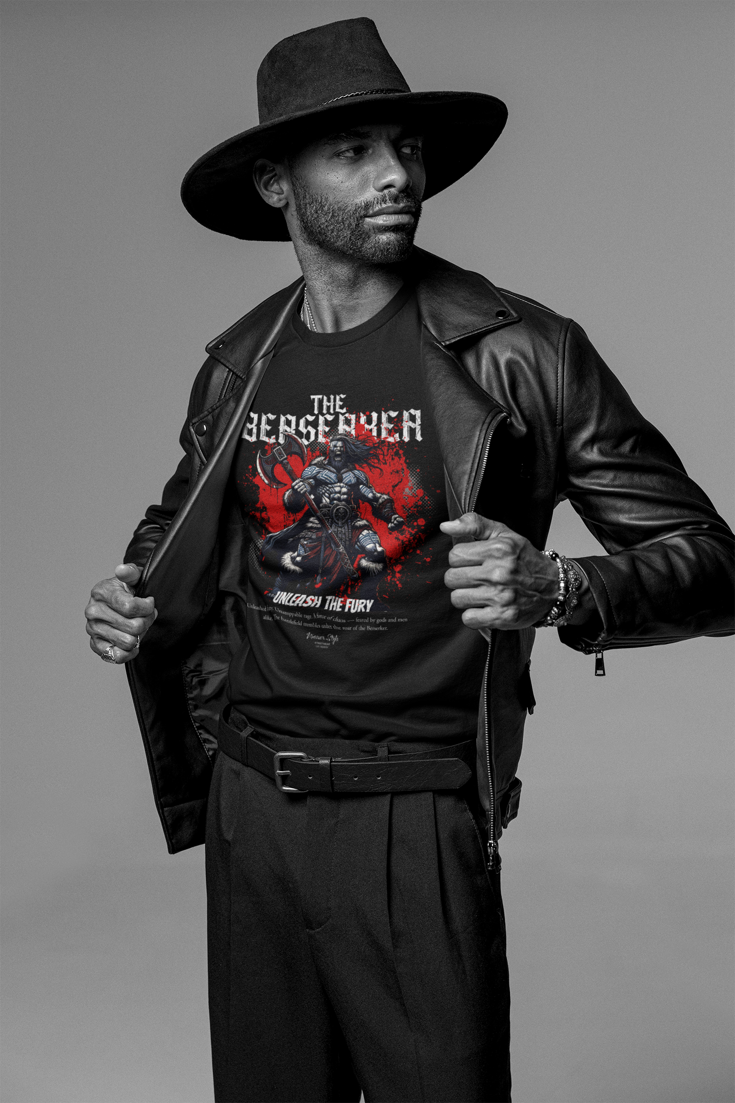 A confident man smiling in a studio shot, opening his leather jacket to reveal THE BERSERKER t-shirt design featuring a battle-worn warrior with twin axes and the slogan “Unleash the Fury”. Monochrome backdrop enhances the dramatic contrast of red and black.