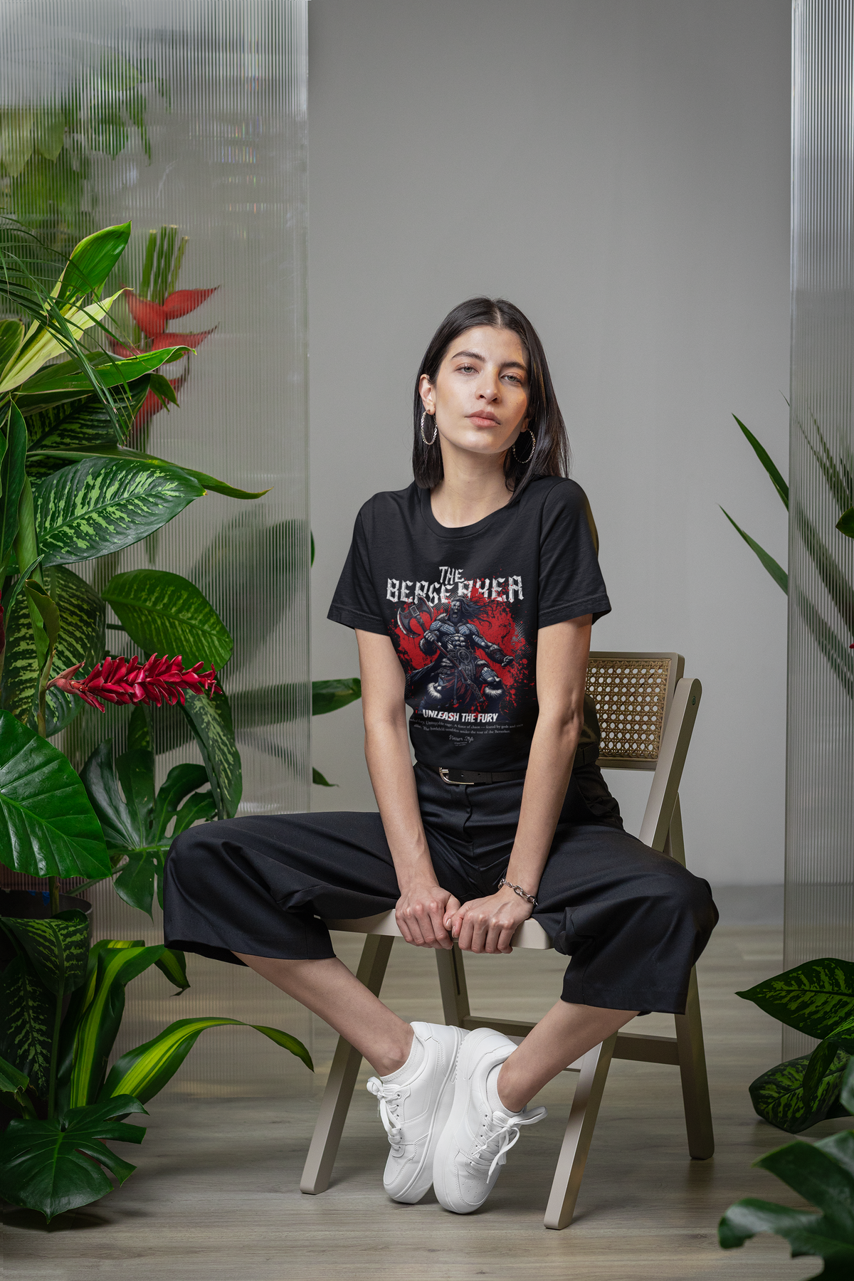 A woman sits confidently among lush indoor plants, wearing THE BERSERKER t-shirt — the fierce Viking design clashing against the calm, natural backdrop. Warrior energy meets botanical stillness.