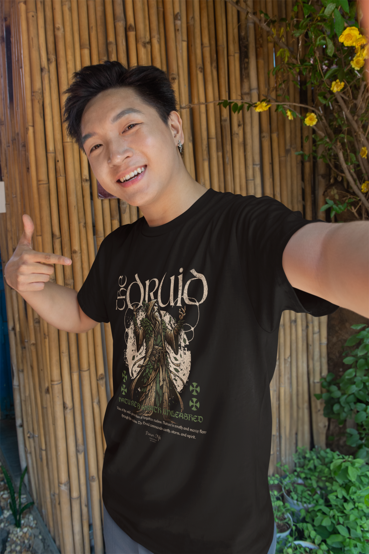 Male model smiling and pointing at his “The Druid” t-shirt in an outdoor bamboo garden setting. Design features a nature mage in forest tones, radiating mystical energy and ancient wisdom.