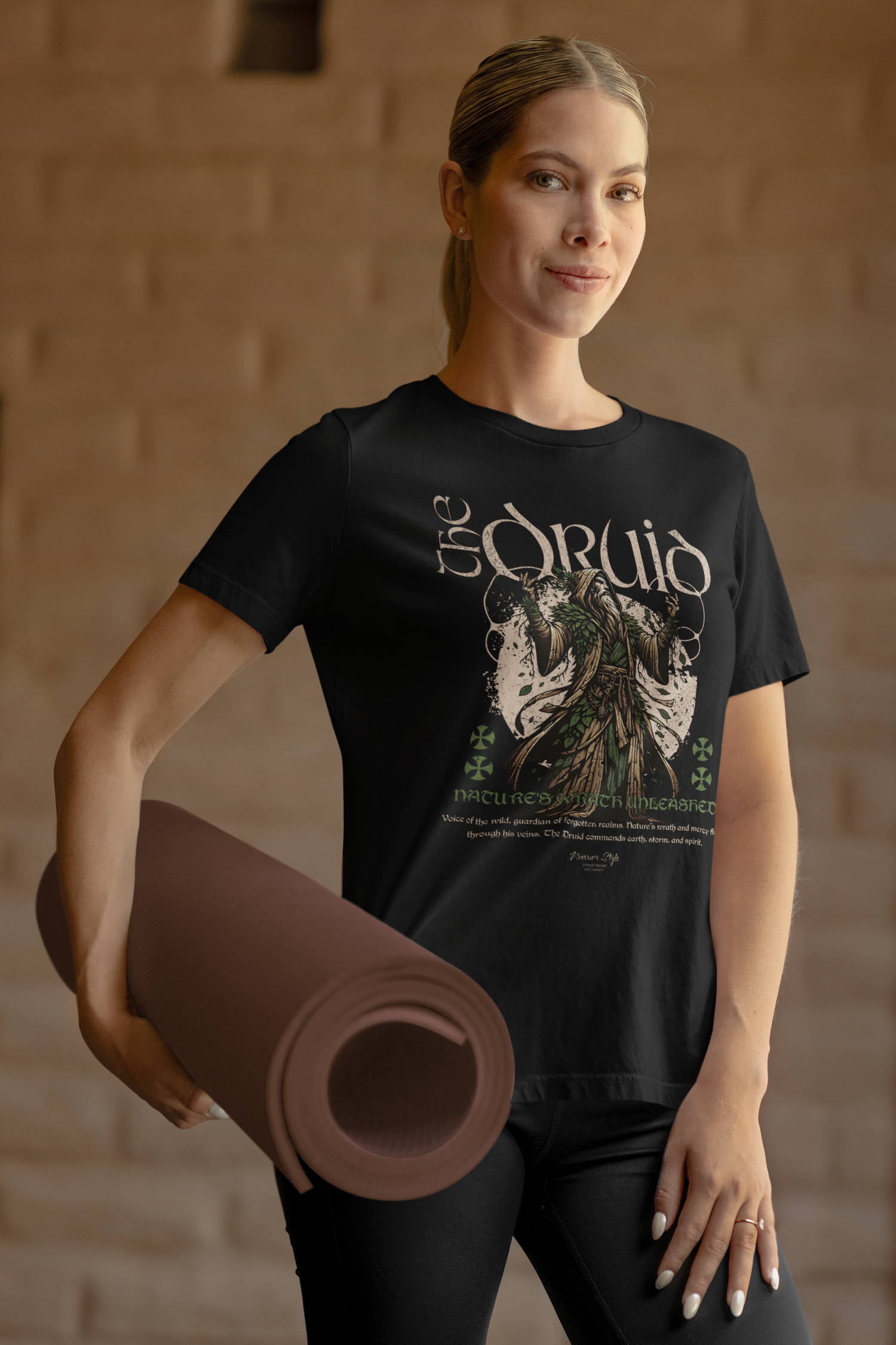 Female model holding a yoga mat in a warmly lit space, wearing “The Druid” t-shirt featuring an ancient forest mage. Earthy tones and spiritual nature theme are clearly highlighted.