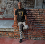 Male model leaning against a brick wall, wearing THE DWARF t-shirt in black — bold golden warrior with dual axes and ‘Forged in Storm and Fire’ tagline.