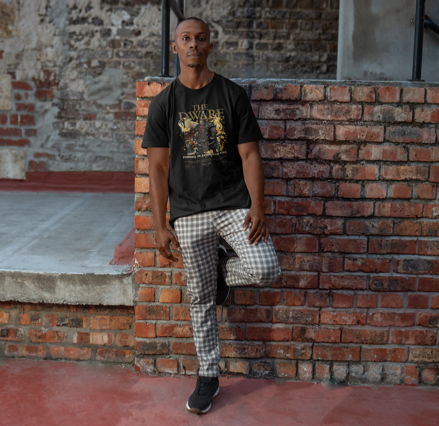 Male model leaning against a brick wall, wearing THE DWARF t-shirt in black — bold golden warrior with dual axes and ‘Forged in Storm and Fire’ tagline.