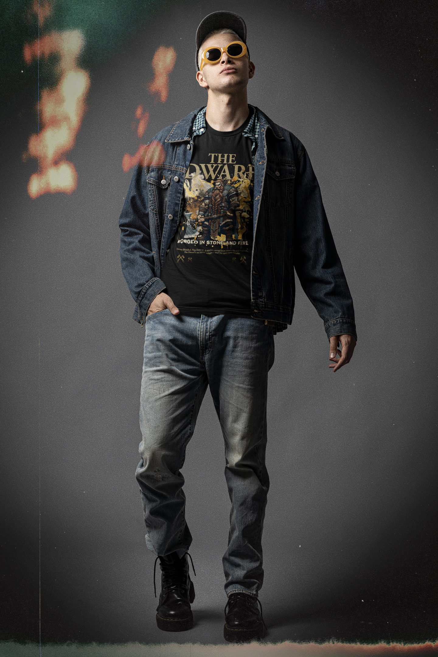 Male model in denim jacket and sunglasses, hands in pockets, wearing THE DWARF t-shirt in black — bold dwarf warrior with axes and golden ‘Forged in Stone and Fire’ design.