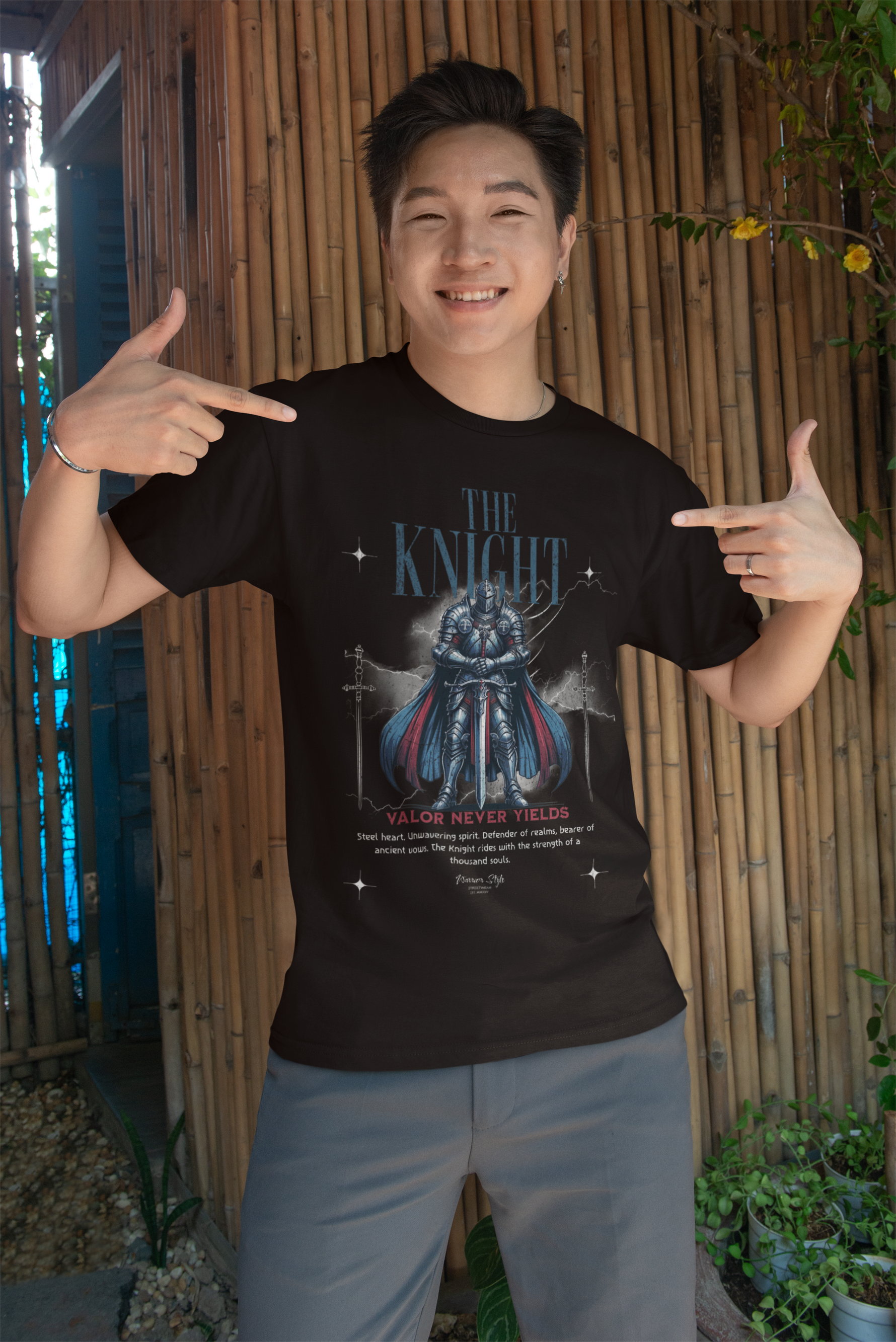 A smiling man proudly pointing at THE KNIGHT t-shirt, featuring a steel-clad warrior and the phrase “Valor Never Yields”, with bamboo walls in the background adding a natural, rustic touch.