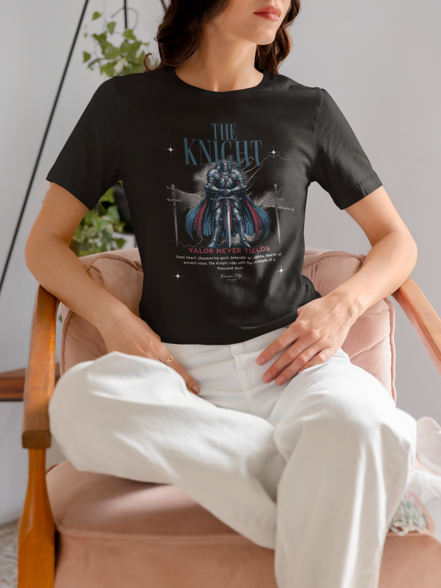 A woman sitting calmly in a velvet chair, wearing THE KNIGHT t-shirt featuring a blue-armored figure and the phrase “Valor Never Yields”, styled with a relaxed yet strong presence.