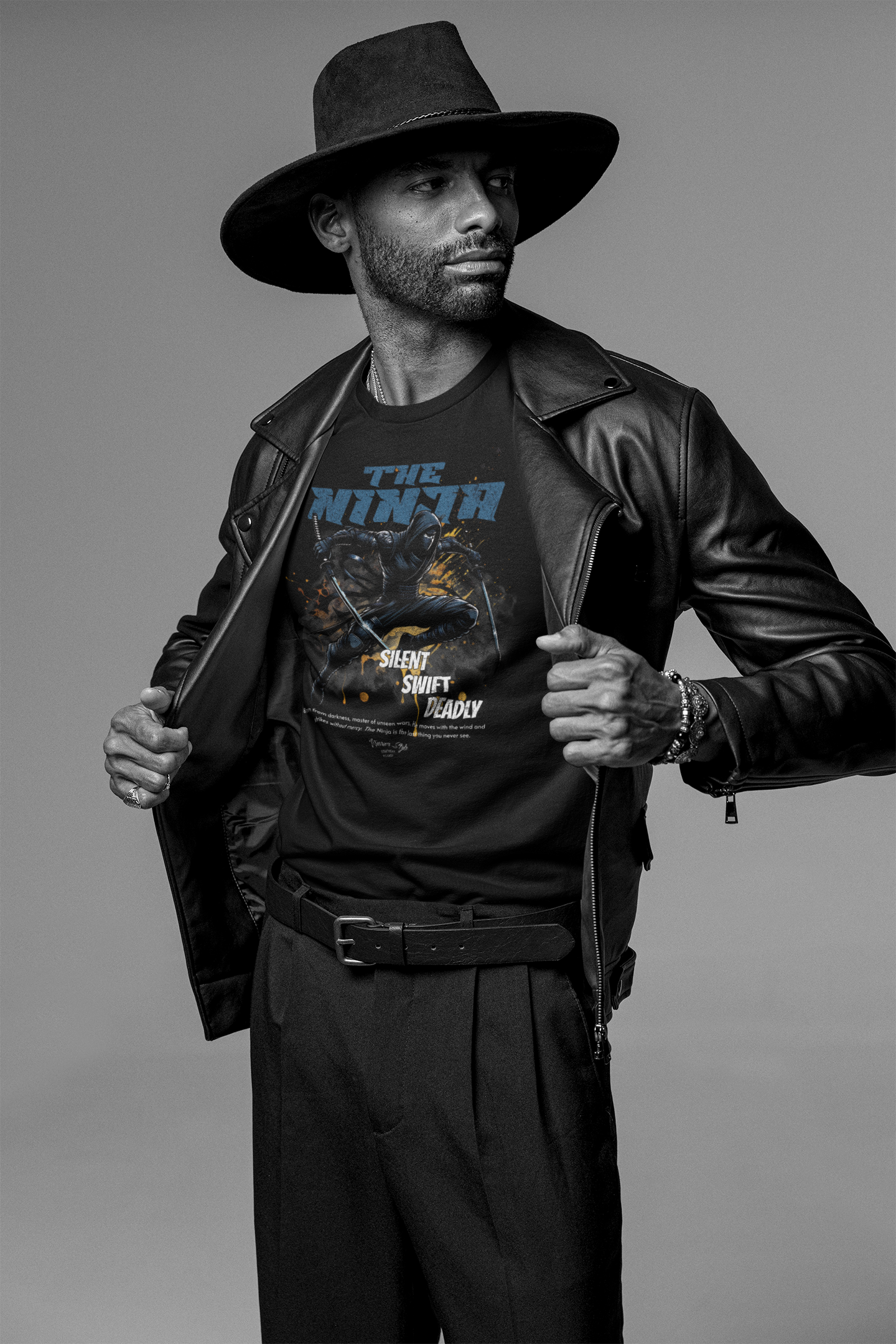 Male model in a wide-brimmed hat and leather jacket revealing the black THE NINJA t-shirt with a stealthy warrior mid-attack pose and bold “Silent. Swift. Deadly.” typography.