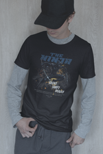 Male model in cap and layered casual outfit, looking down while wearing the black THE NINJA t-shirt featuring a crouching ninja and the bold phrase “Silent. Swift. Deadly.”