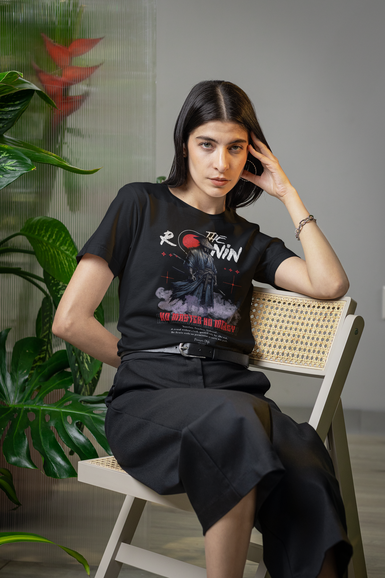 Female model sits relaxed on a wooden chair, wearing The Ronin tee — lone warrior design boldly stands out amidst indoor plants.