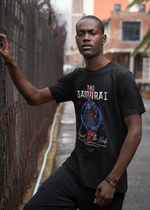 A confident man standing in an urban alley, gripping a rusted fence with one hand, wearing THE SAMURAI t-shirt design featuring a red-blue armored warrior and the slogan “Honor Is The Blade”. Weathered bricks and industrial structures frame the gritty background.