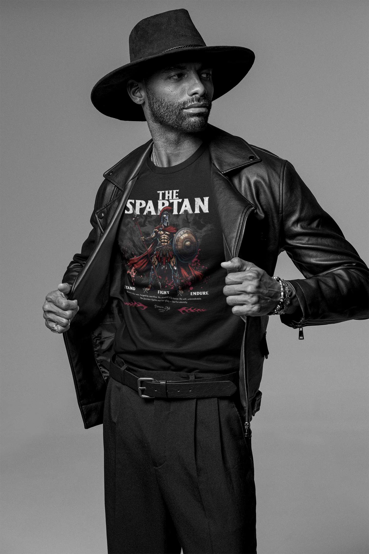 A bold male model in a black leather jacket and wide-brim hat reveals THE SPARTAN t-shirt. The design features a warrior with shield and spear, captioned with “Stand. Fight. Endure.” in red and white.