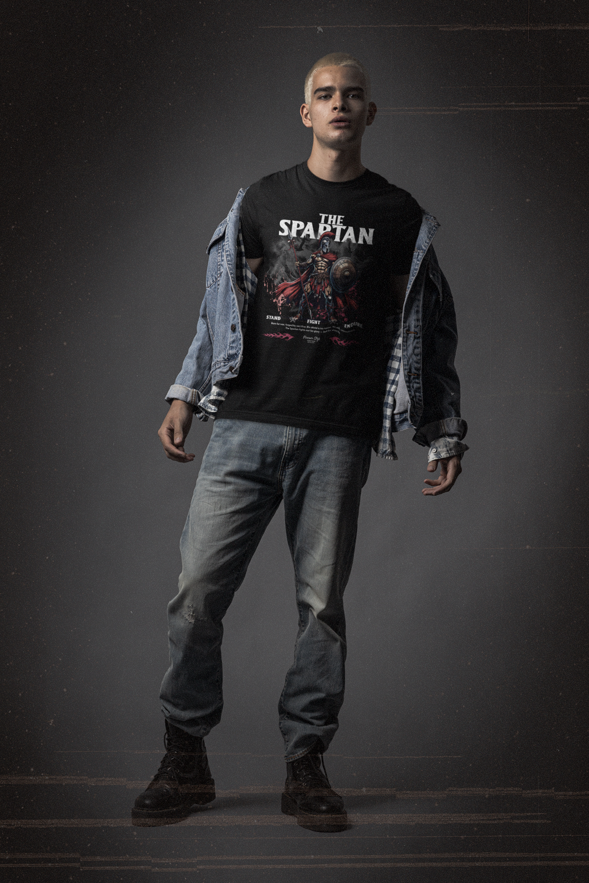 A bold male model in denim and boots stands strong in THE SPARTAN t-shirt. The design features a battle-worn Spartan warrior with the words “Stand. Fight. Endure.” beneath him.