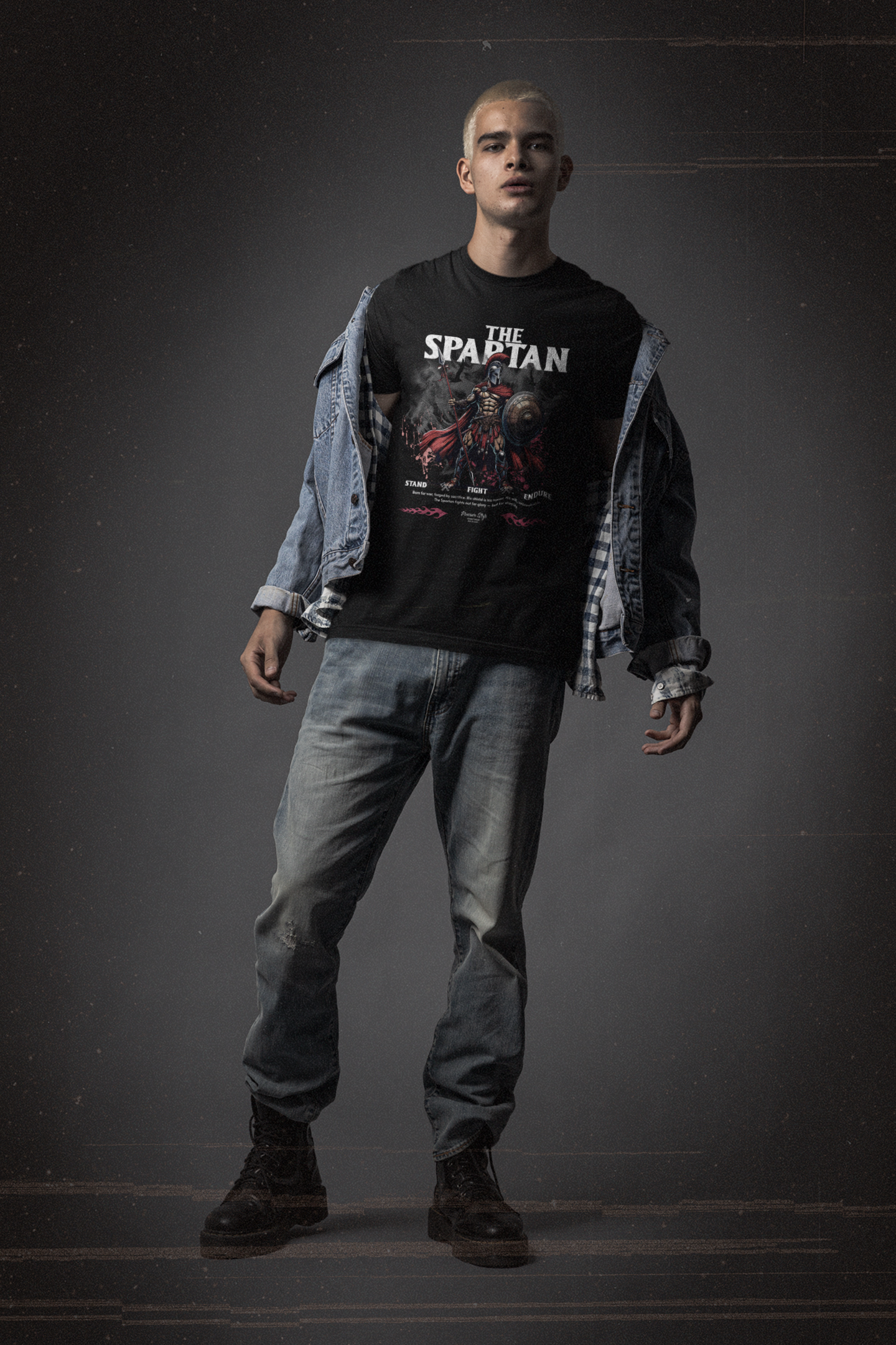 A bold male model in denim and boots stands strong in THE SPARTAN t-shirt. The design features a battle-worn Spartan warrior with the words “Stand. Fight. Endure.” beneath him.