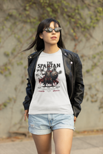 A confident female model in a black leather jacket and shorts walks outdoors wearing THE SPARTAN t-shirt. The design shows a Spartan warrior with blood-red accents and the quote “Stand. Fight. Endure.”