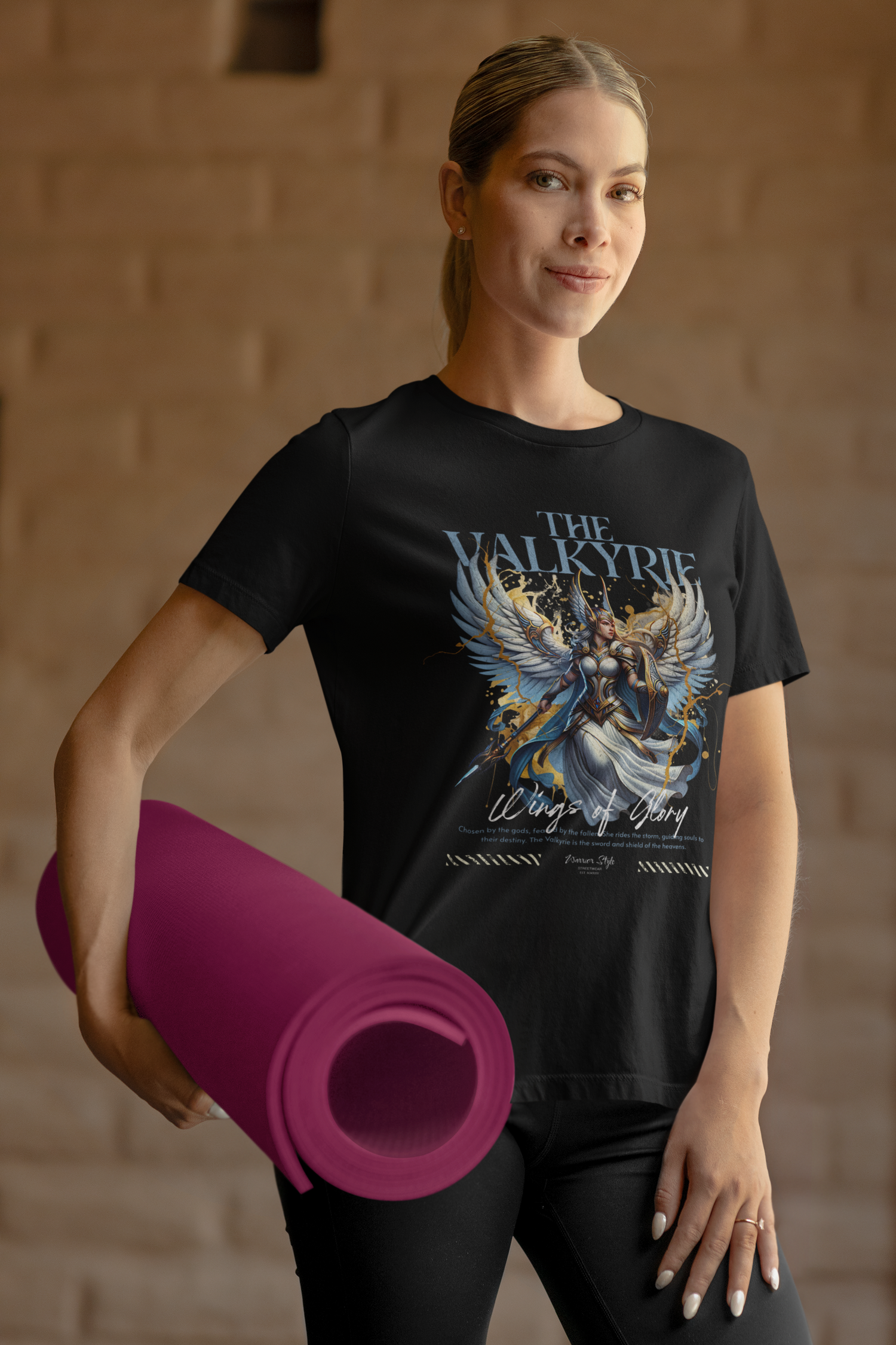 A calm female model holding a rolled yoga mat stands in soft indoor light, wearing THE VALKYRIE t-shirt. The artwork features a winged warrior with golden accents and the phrase “Wings of Glory.”