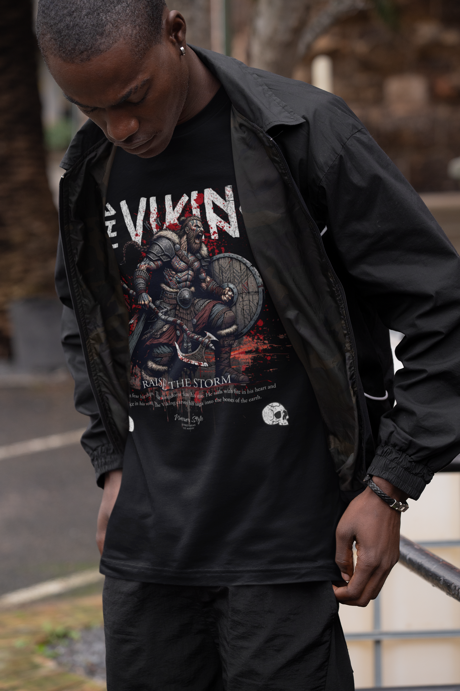 A man looking down with intensity, standing by an iron railing in a courtyard, revealing THE VIKING t-shirt design with a raging Norse warrior and the slogan “Raise The Storm”.