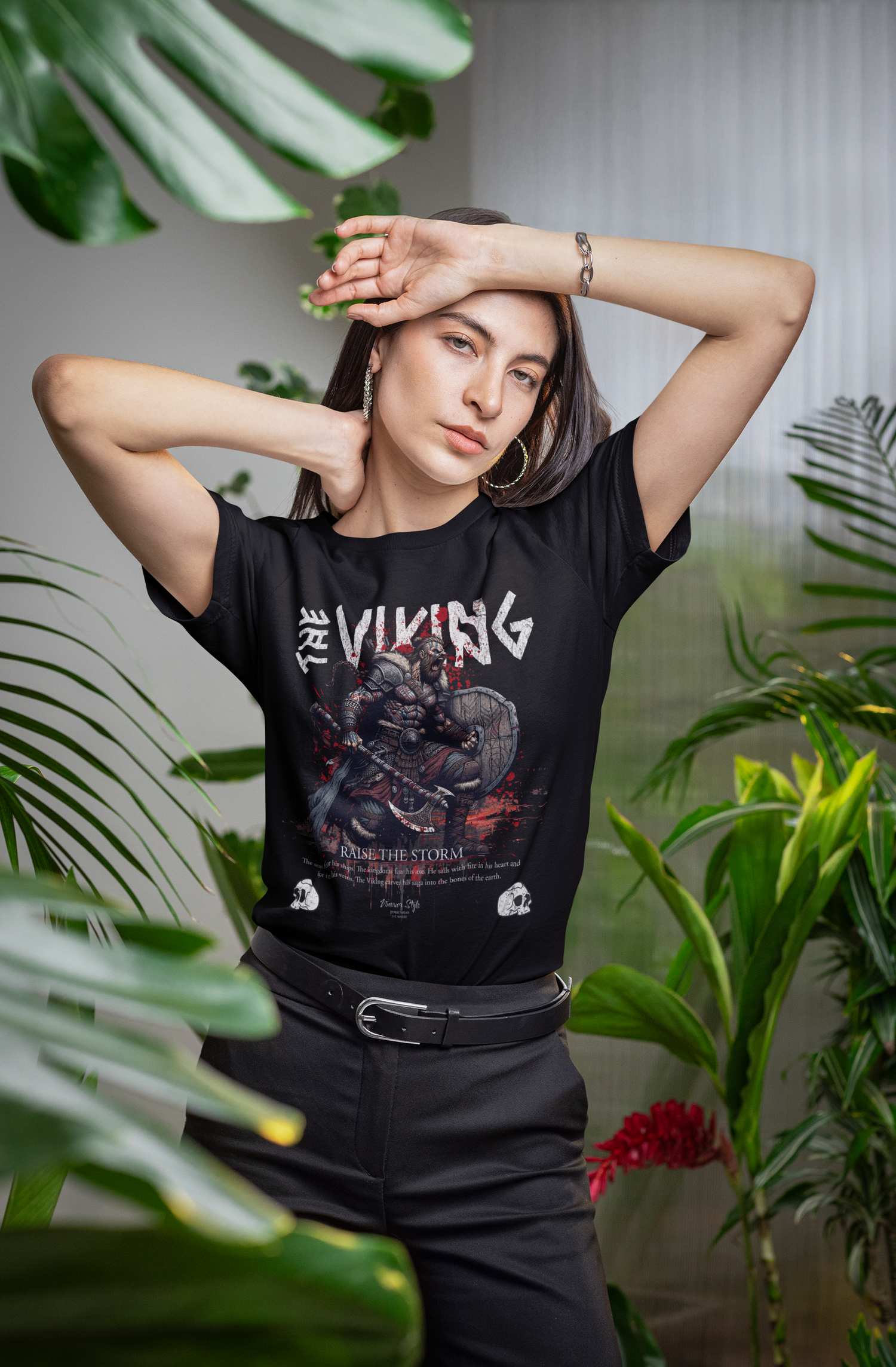 A woman striking a bold pose among lush indoor plants, wearing THE VIKING t-shirt featuring a fierce Norse warrior and the slogan “Raise The Storm”.
