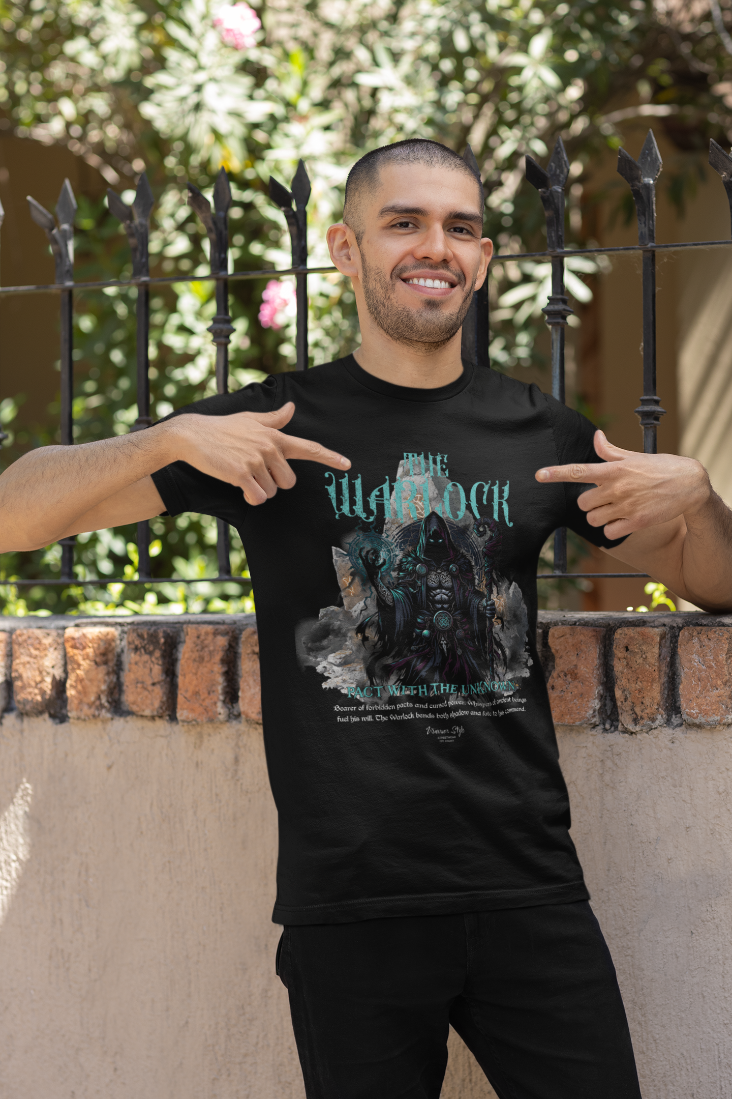 A confident man smiles while pointing at his black THE WARLOCK t-shirt — embracing arcane power under the midday sun.