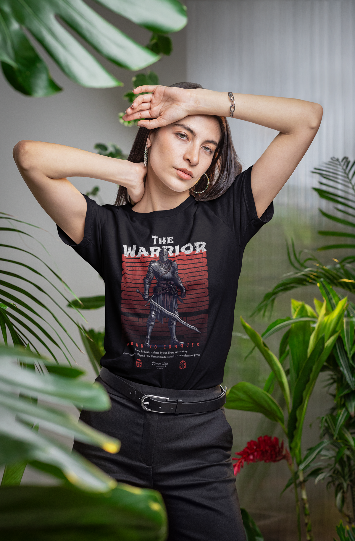 Woman in tropical indoor setting wearing THE WARRIOR t-shirt — striking a bold pose among lush plants, highlighting the medieval knight graphic and 'Born to Conquer' text.