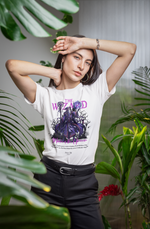 Confident woman posing among indoor plants, wearing THE WIZARD t-shirt. Her arms frame her face dramatically, highlighting the electric purple design of the wizard casting lightning on the shirt.