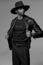 A confident man wearing a black leather jacket and wide-brimmed hat reveals the black TOKYO t-shirt underneath — featuring a minimalist city badge with Tokyo Tower, 5-star layout, and a red-white progress bar. Monochrome studio background adds a timeless, cinematic feel.